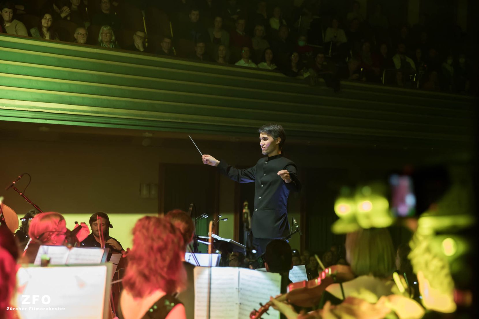 Ein Dirigent leitet sein Orchester, von dem ein Ausschnitt zu sehen ist. Im Hintergrund die Zuhörer im Zürcher Volkshaus.